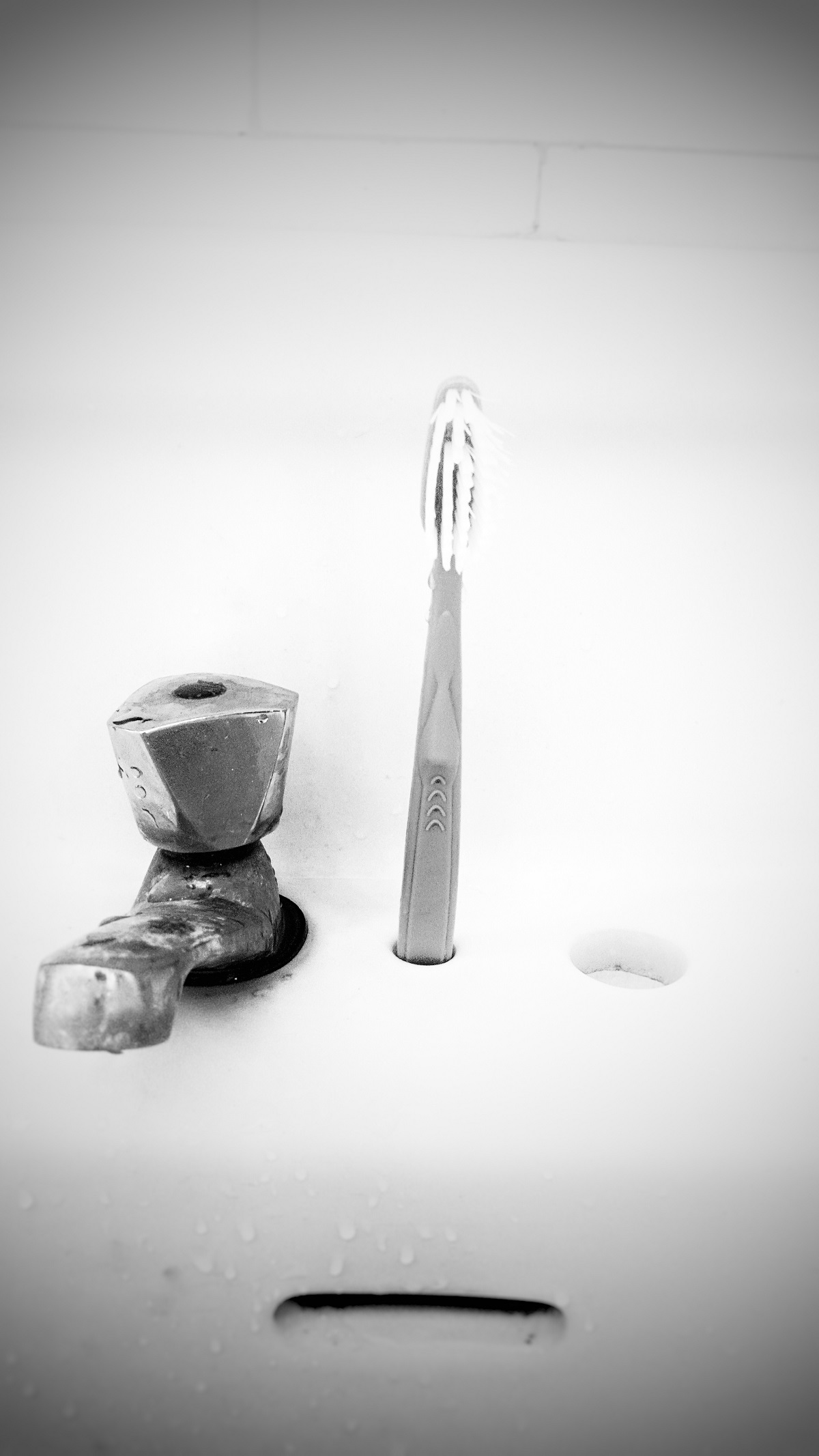 Photo noir et blanc. Elle montre un lavabo sur lequel une brosse à dent est positionnée à la verticale, à droite du robinet.