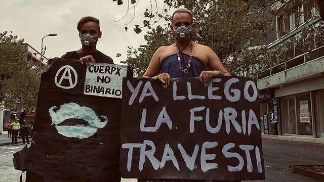 Deux personnes tiennent des pancartes écrites en blanc sur fond noir, et portent des masques sur le visage lors de la marche nationale trans en Equateur.