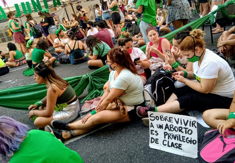 Un groupe de jeunes filles portant des accessoires verts, sont assisses par terre et portent des affiches en faveur de l'avortement sûr.