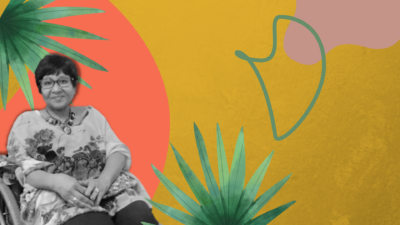 Cristina Franco está sentada em sua cadeira de rodas. Ela tem o cabelo curto e usa óculos, anéis em ambas as mãos e um colar de pedras. Veste uma blusa longa, estampada com desenhos florais. A foto tem o fundo ilustrado com folhas de palmeira caribenha.