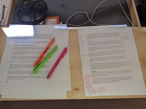 White sheets of printer-sized paper are laid out on a desk, they contain the articles for this reporting project. The papers are covered with handwritten annotations in green, pink and orange pen. 