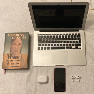 The keyboard of Lida's laptop has some keys used so much that the letters have been handwritten on again. The laptop sits beside her cell phone, her wireless airpods, and a print book titled "Malena is a Name of a Tango" by author Almudena Grandes.