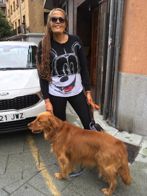 Lida Margarita Carriazo has long, brown, braided hair and wears sunglasses and a long T-shirt with a Mickey Mouse design. She stands on a street in Spain with her guide dog Raquel, a Golden Retriever.