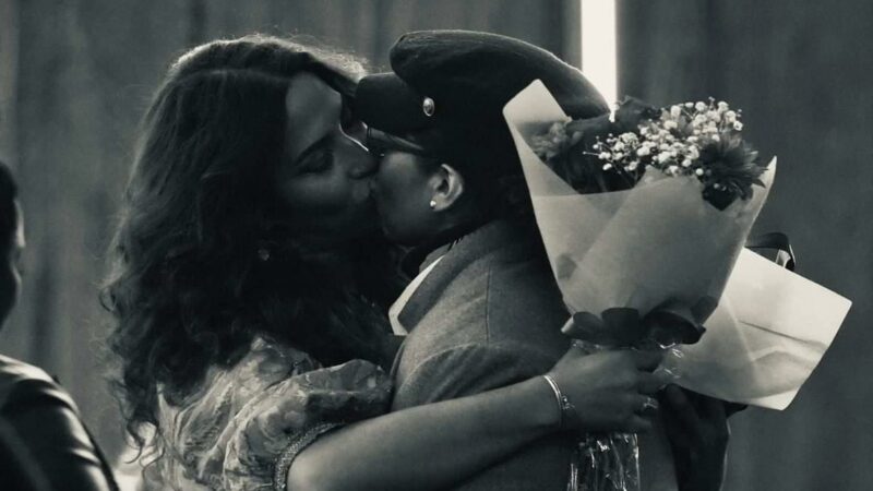 Une femme aux cheveux longs et une autre avec une gavroche s'embrassent, un bouquet de fleurs à la main