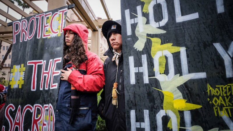 deux femmes avec des vestes de pluie à côté panneaux de protestation plus grands qu'elles, l'une des deux à la main sur le cœur et les yeux fermés