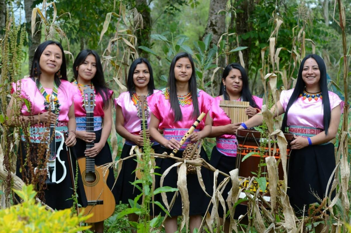 Six femmes, en tenue traditionnelle rose et noire, posent dans la nature, avec leur instrument de musique respectif : violon, guitare, charango, flute, flute de pan et tambour