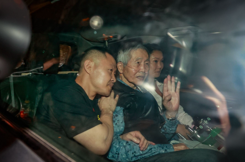 À travers une vitre de voiture, un homme âgé au centre, portant une canule nasale et saluant de la main, entouré d'un homme et d'une femme